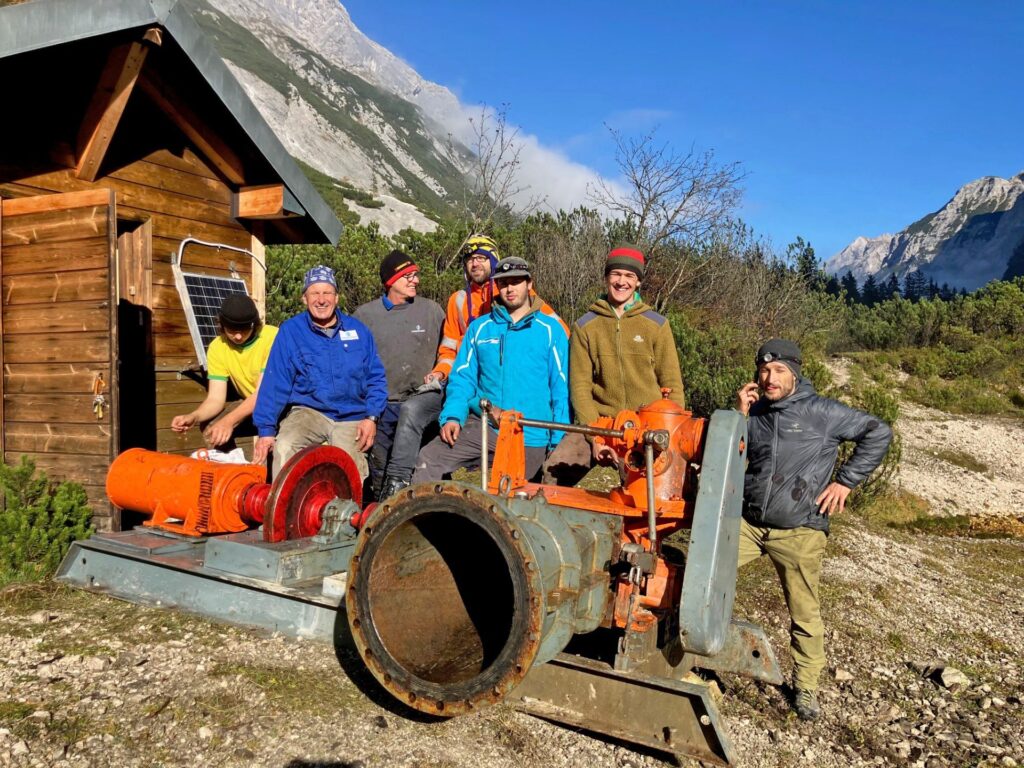 Siebenköpfiges Team bei Montagearbeiten an einer großen Industrie-Wasserpumpe vor einer Almhütte im Hochgebirge.