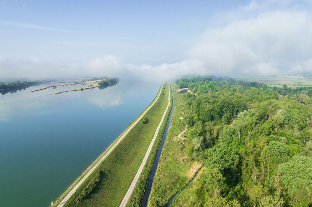 Luftaufnahme eines blauen Flusses neben einem grünen Damm mit Radweg, angrenzender Wald und Nebel am Horizont.