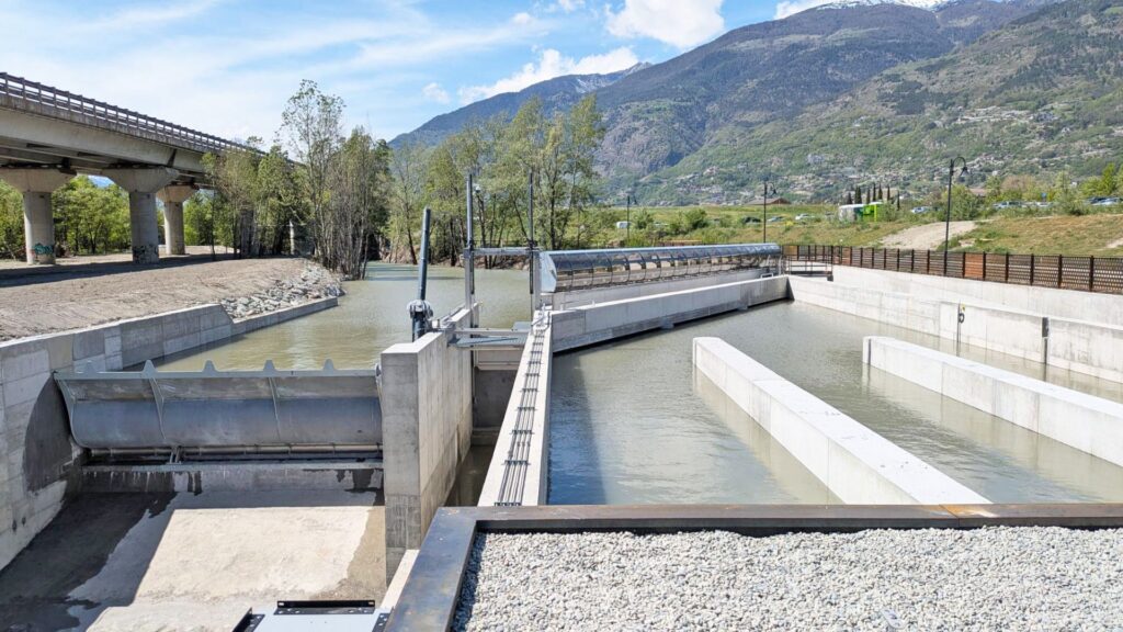 Wasserbauliche Anlage in Aosta mit Betonschleusen und Wehr vor bewaldeten Bergen unter blauem Himmel.