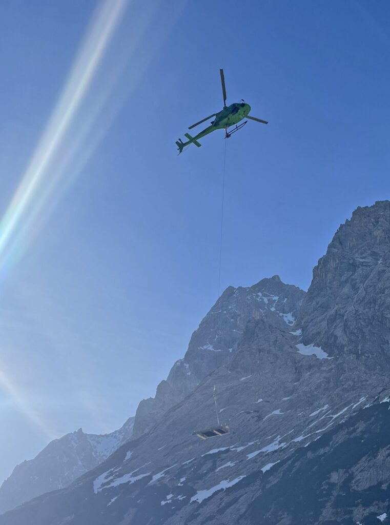 Grüner Hubschrauber beim Helitransport einer Last am Seil vor schneebedeckten Berggipfeln und blauem Himmel.