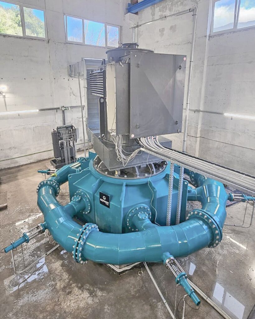 Große Mega Rema Wasserturbine von Geppert mit blauen Rohrleitungen und grauem Generatorgehäuse in einer Industriehalle.