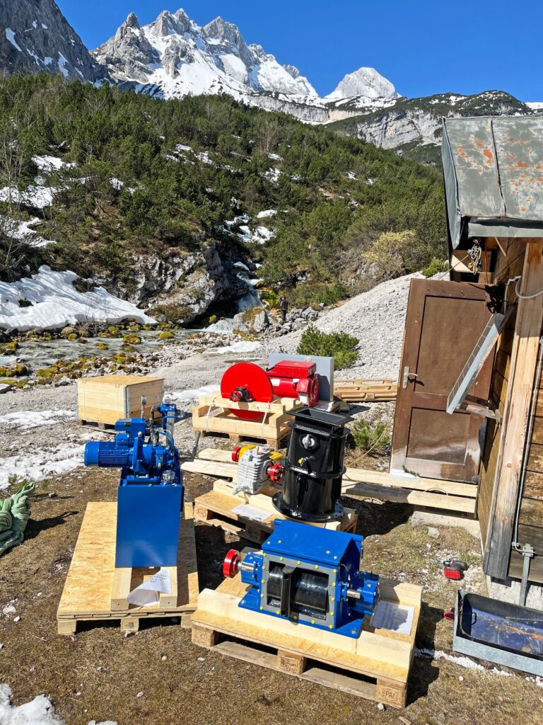 Fünf Neuteile für das Turbinenhaus auf Paletten vor verschneitem Hochgebirge am Bergbach.