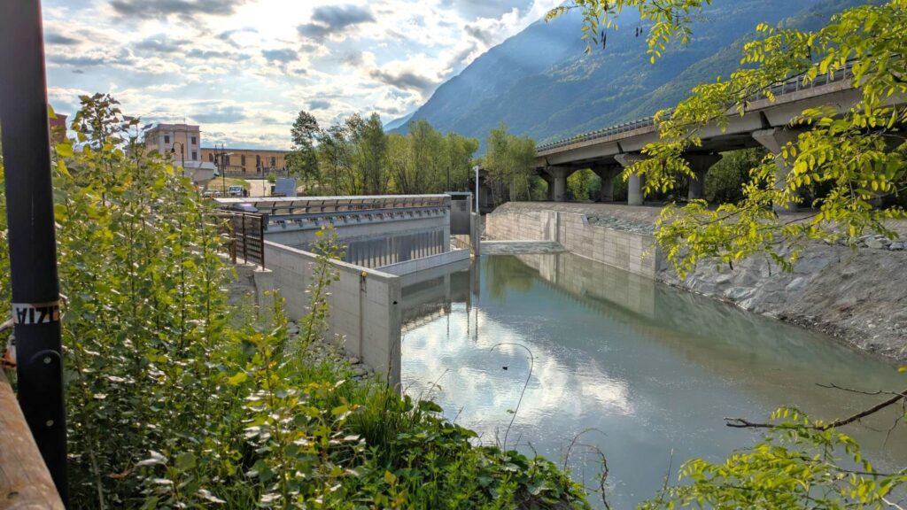 Stauanlage im Aostatal vor Alpenpanorama mit Autobahnbrücke, Betonmauern und ruhigem Flusslauf unter bewölktem Himmel.
