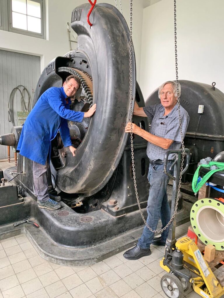 Zwei Techniker arbeiten an einer großen, gusseisernen Wasserturbine in einer historischen Maschinenhalle.
