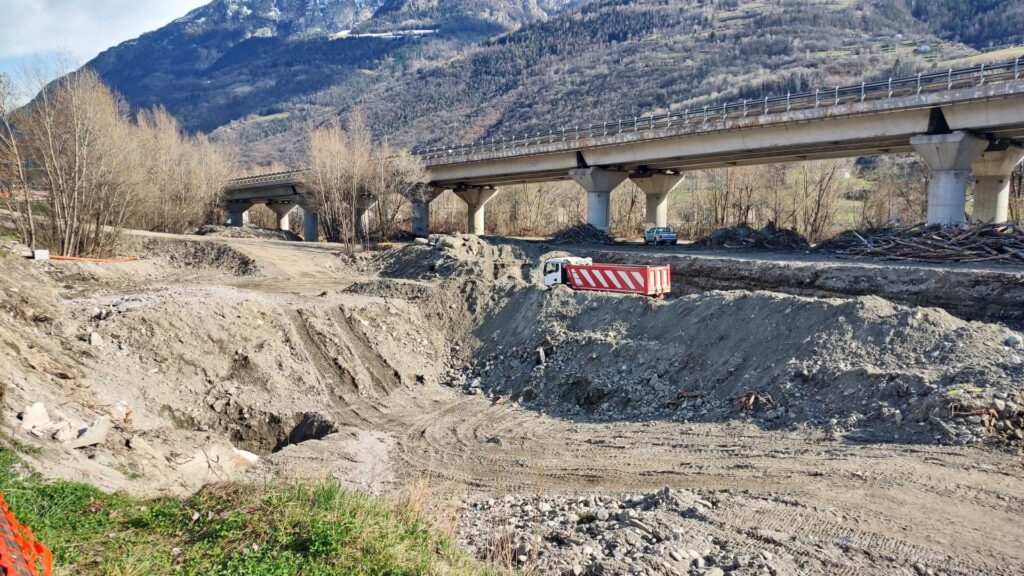 Baubeginn mit Erdarbeiten und LKW vor einer Autobahnbrücke in alpiner Landschaft mit Bergen im Hintergrund.