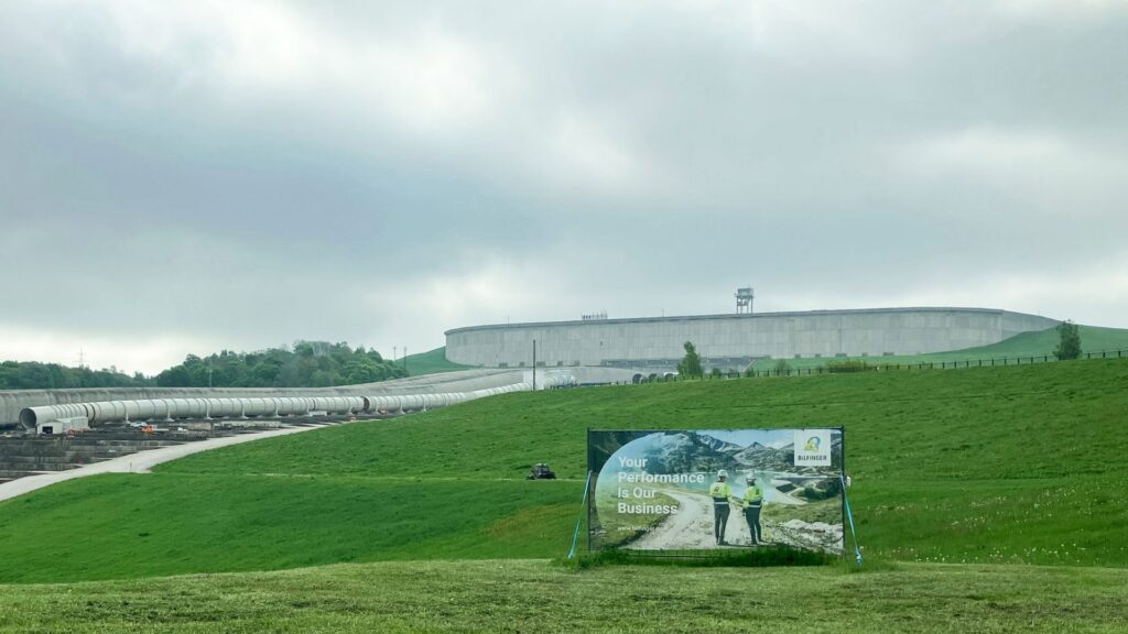 Bilfinger Werbebanner auf grüner Wiese vor Industrieanlage mit Rohrleitungen und Betonbauwerk unter bewölktem Himmel.