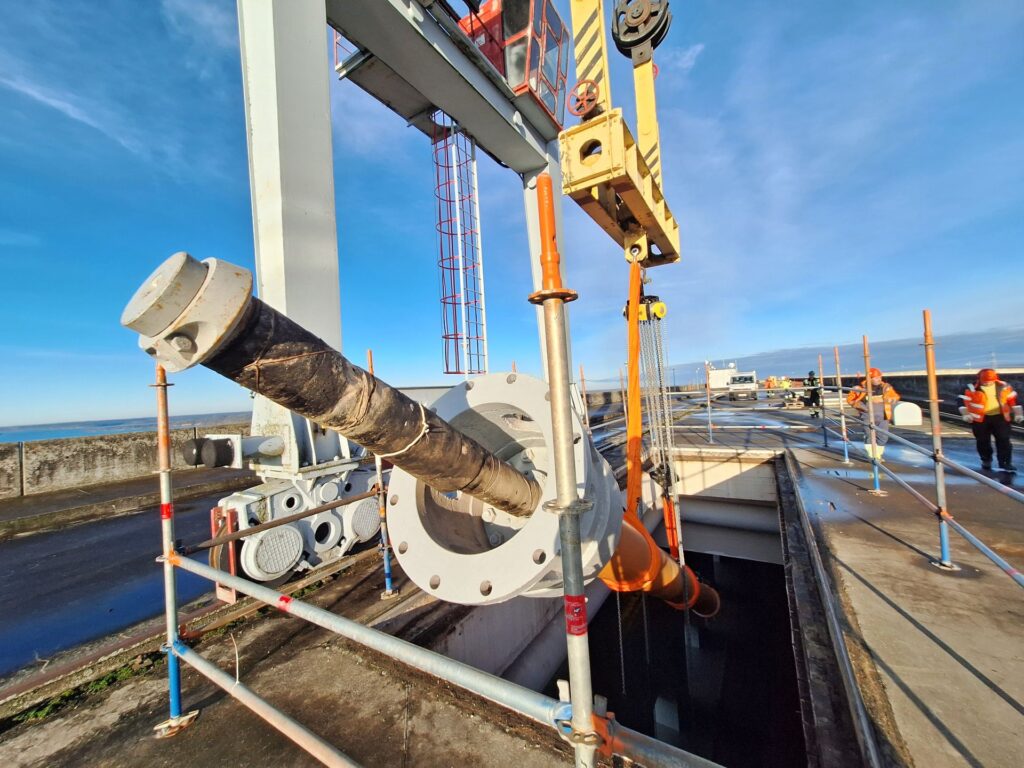 Portalkran hebt massives Rohrbauteil auf einer Baustelle mit Arbeitern in Warnweste bei strahlendem Sonnenschein.