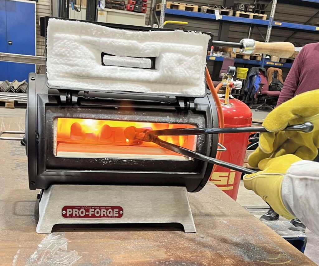 Geöffneter Pro-Forge Gassedeschmiedeofen mit glühendem Innenraum; eine Zange hält Metall im Feuer eines Brennofens.