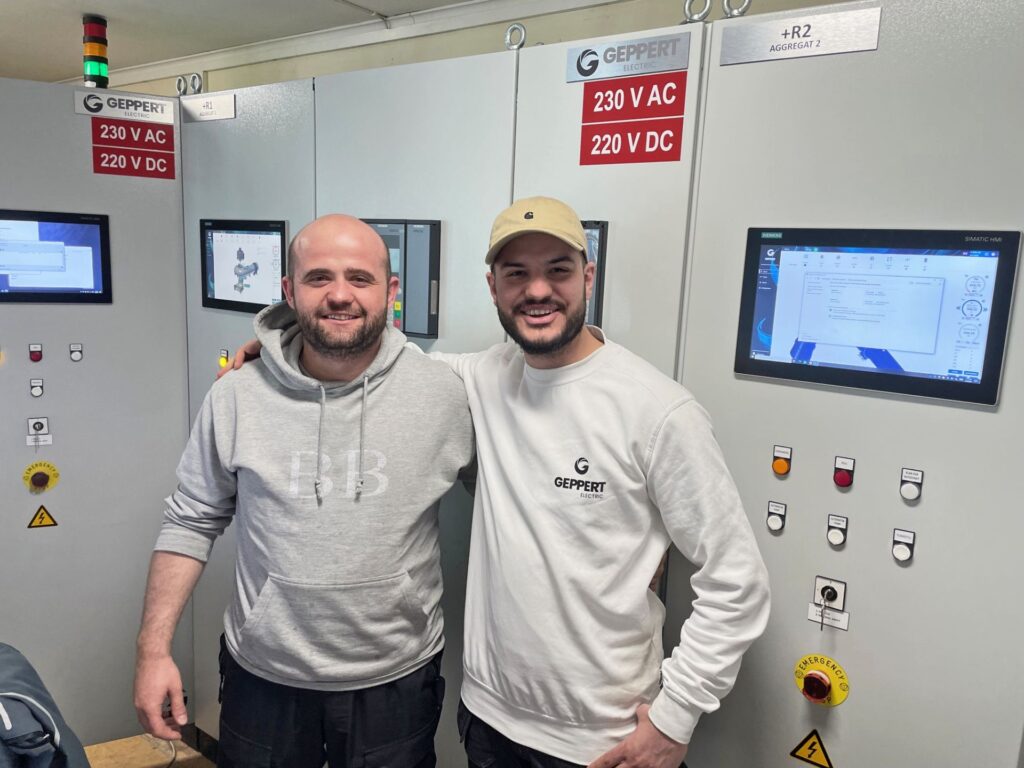 Zwei lächelnde Techniker von Geppert Electric vor einem industriellen Control System Schaltschrank mit Displays.