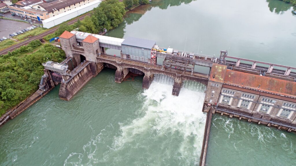 Luftaufnahme eines Flusskraftwerks mit Staustufe, Wehranlage und turbulent abfließendem Wasser im grünen Flussbett.