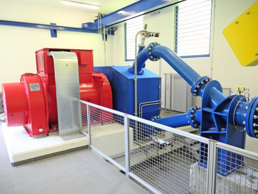 Industrieller AEM Generator in Rot neben einer blauen Wasserturbine mit Rohrleitungen in einem Maschinenraum.