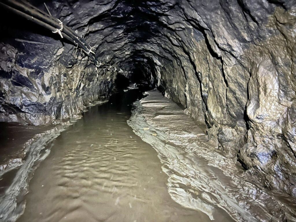 Unterirdischer Druckstollen mit felsigen Wänden, Wasser am Boden und Kabeln an der Decke, die in die Dunkelheit führen.