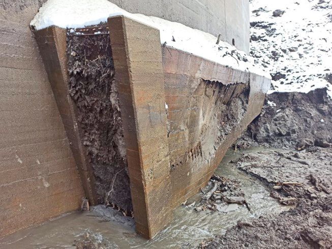 Beschädigtes Einlaufbauwerk aus Beton mit Rissen und Ausspülungen in einer verschlammten Winterlandschaft.