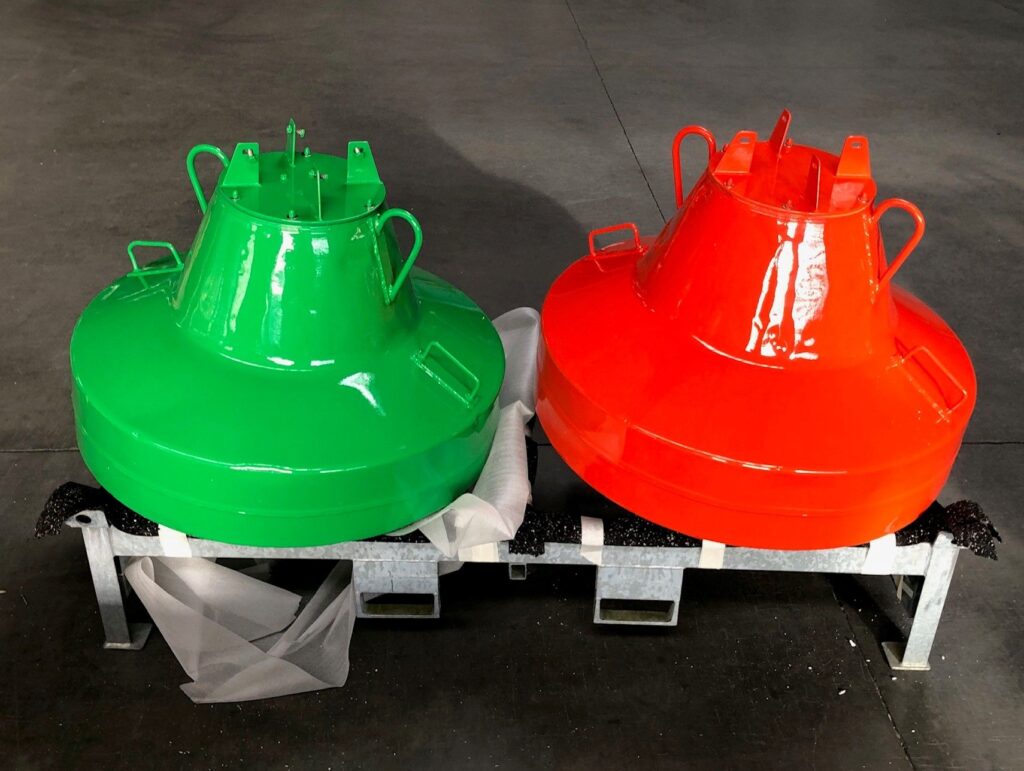 Grüne und rote Fahrwassertonnen aus Metall nebeneinander auf einem Gestell in einer Werkshalle.