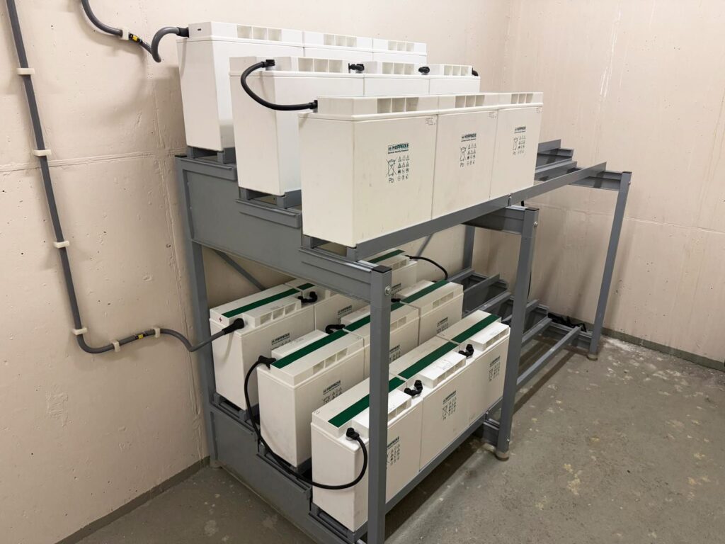 Neu installierte 220VDC Batterien von Hoppecke auf einem grauen Metallregal in einem Technikraum.