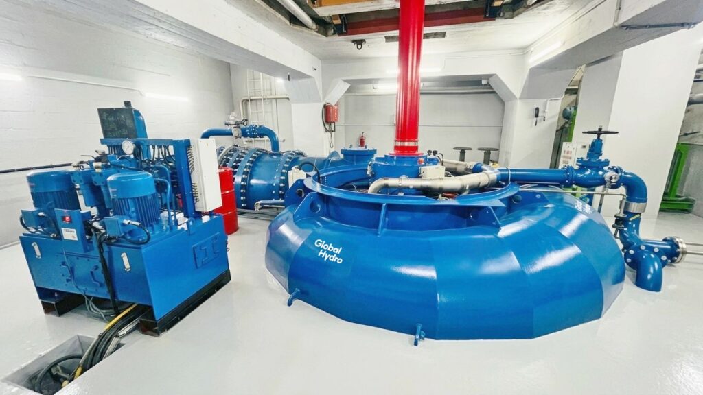 Blaue Global Hydro Turbine im Powerhouse mit rotem Steigrohr und strukturierter Kabelführung am Boden.