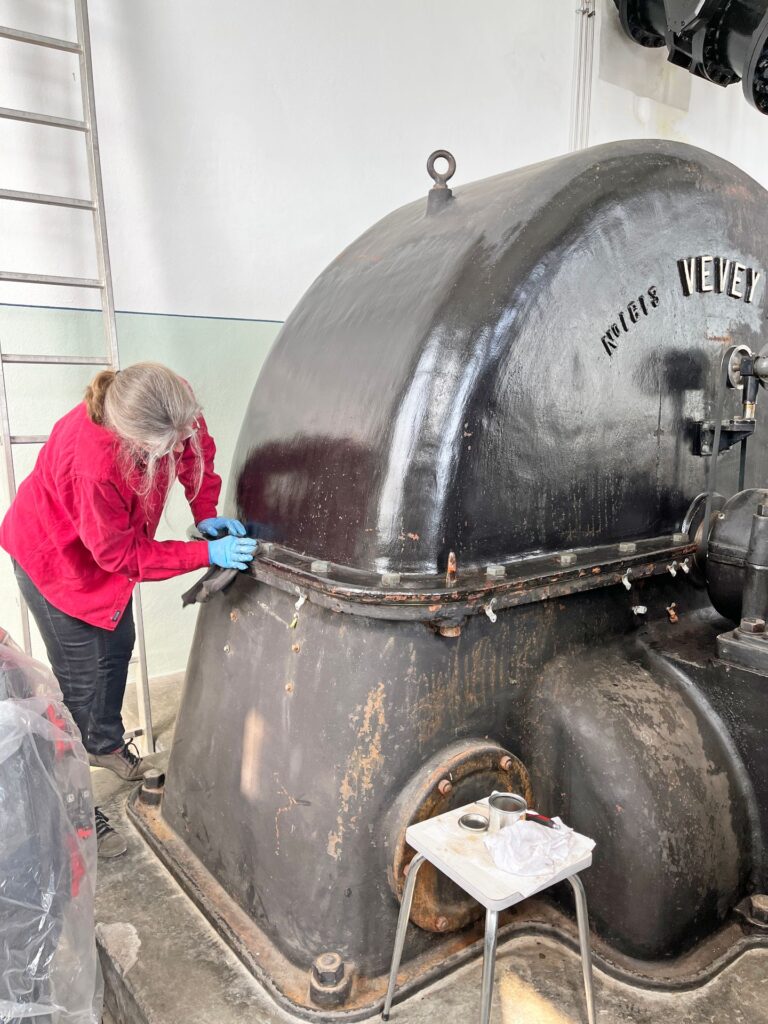 Restaurierung einer historischen Ateliers Vevey Wasserturbine durch eine Frau in roter Jacke mit Schutzhandschuhen.