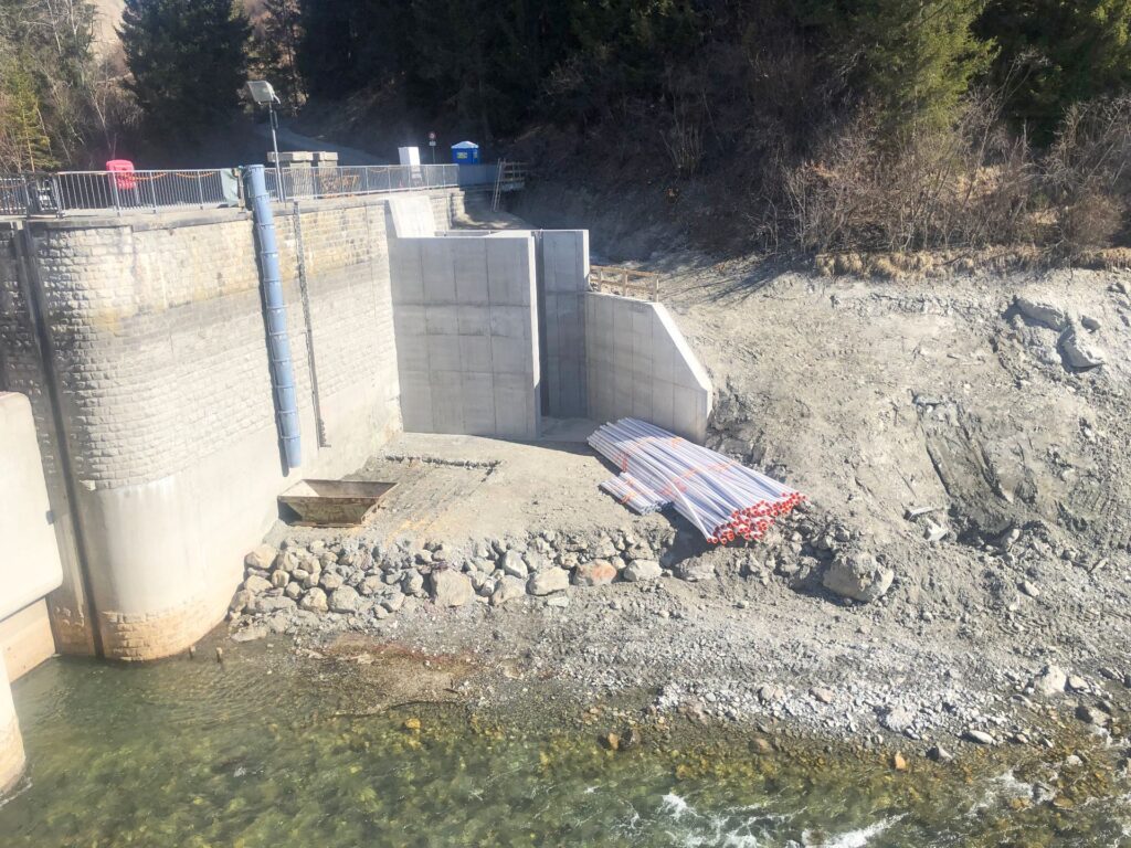 Baustelle an einer Staumauer mit Betonwänden, Rohren und Steinen am Flussufer unterhalb eines bewaldeten Hangs.