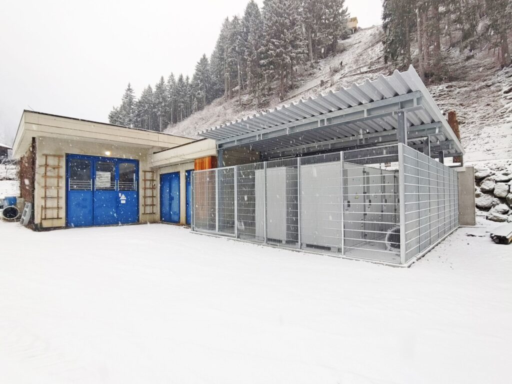 Eingezäunte technische Anlage mit Wellblechdach neben Gebäude mit blauen Toren im verschneiten Gebirge vor Nadelwald.