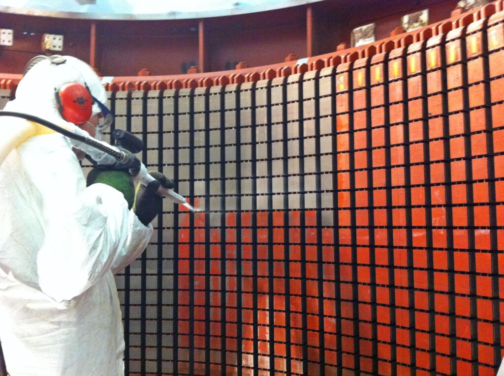 Arbeiter in Schutzkleidung führt Trockeneisreinigung an einem großen orangefarbenen Generator-Stator durch.
