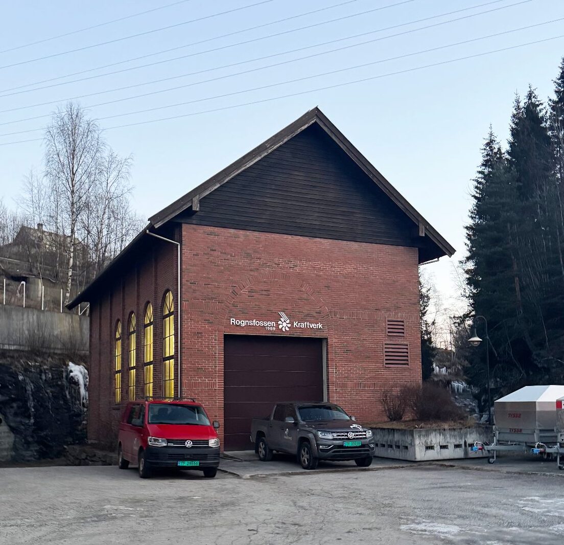 Backsteingebäude des Rognsfossen Kraftwerk mit parkenden VW-Fahrzeugen vor dem Garagentor und bewaldetem Hintergrund.