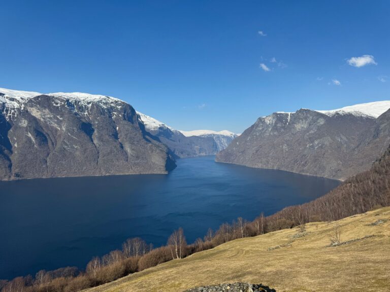 Panoramablick auf den tiefblauen Aurlandsfjord in Norwegen zwischen steilen, schneebedeckten Bergen unter klarem Himmel.
