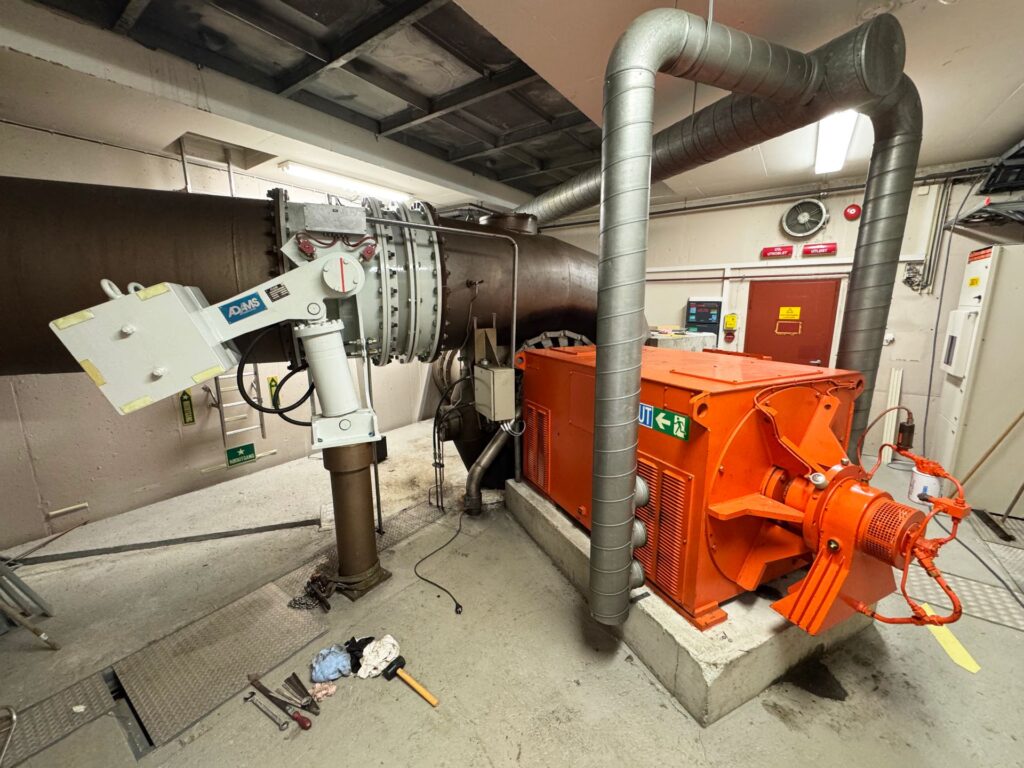 Großer oranger Industriemotor mit massiven Rohrleitungen und Adams-Hydraulikarm in einem technischen Versorgungsraum.