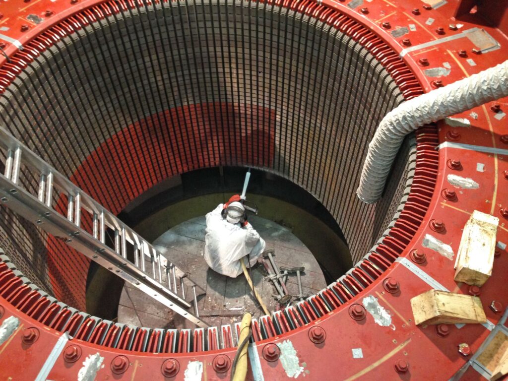 Industriearbeiter im Schutzanzug bei der Wartung im Inneren eines großen, roten Generator-Stators in einem Kraftwerk.