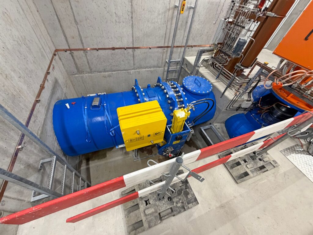 Große blaue Industrie-Rohrleitung mit gelber Steuereinheit in einem Betonschacht einer technischen Anlage.