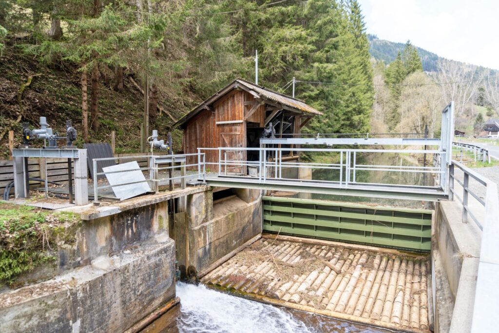 Wehranlage des Kraftwerks Hüttenberg mit Holzhaus, grünem Wehrschütz und Rechenreiniger vor bewaldetem Hang.