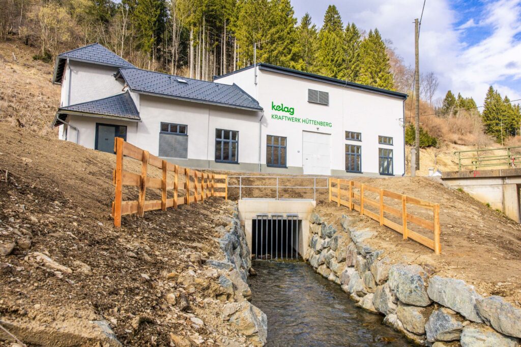 Außenansicht Kelag Kraftwerk Hüttenberg mit Wasserlauf, Rechen und Steineinfassung vor bewaldetem Hang.