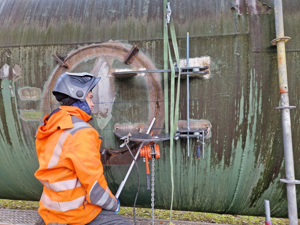 Arbeiter mit Schweißhelm montiert Vorrichtung an einem Mannloch eines großen, grünen Metalltanks im Außenbereich.