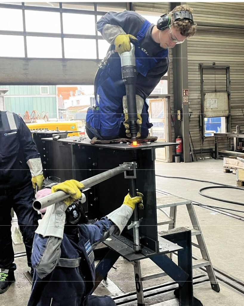 Industrieller Nietprozess an einem Stahlträger mit glühendem Niet und zwei Facharbeitern in einer Werkstatthalle.