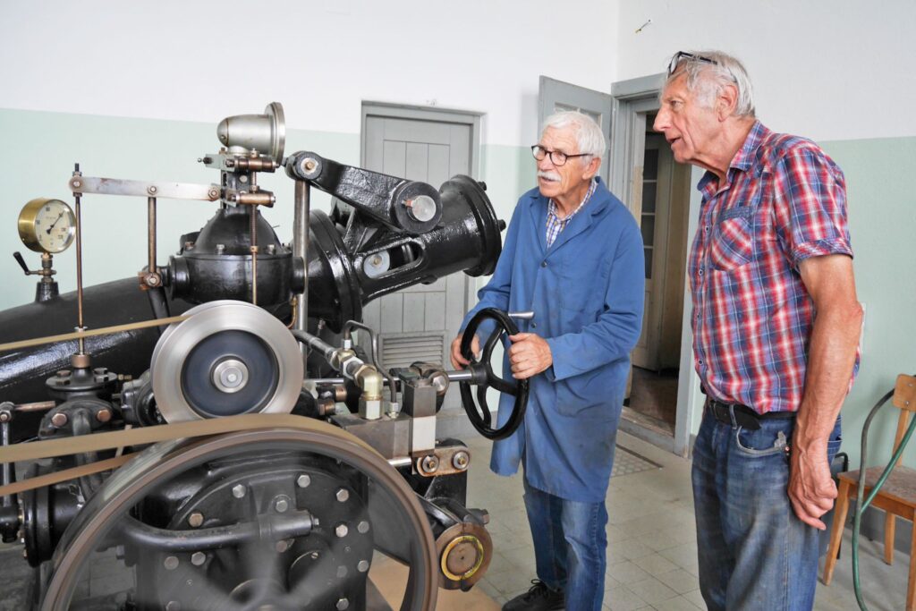 Zwei ältere Männer in Werkstattkleidung bedienen eine historische, schwarze Industriemaschine mit Antriebsriemen.