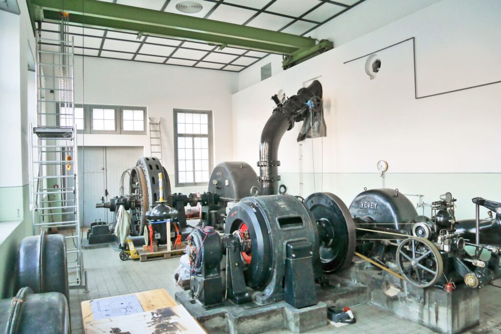 Historische Vevey-Wasserturbinen und Generatoren in einer Industriehalle mit Stahlrohren, Transmissionsrädern und Leiter.