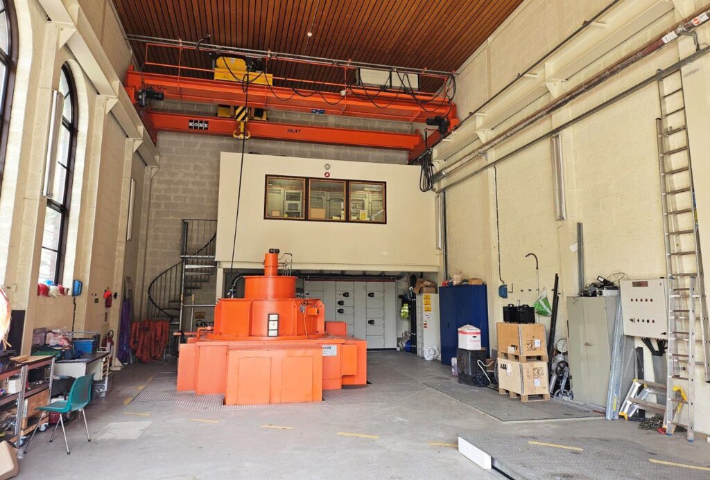 Innenansicht eines Powerhouse mit orangefarbener Industrieturbine, Deckenkran, Kontrollraum und technischer Ausrüstung.
