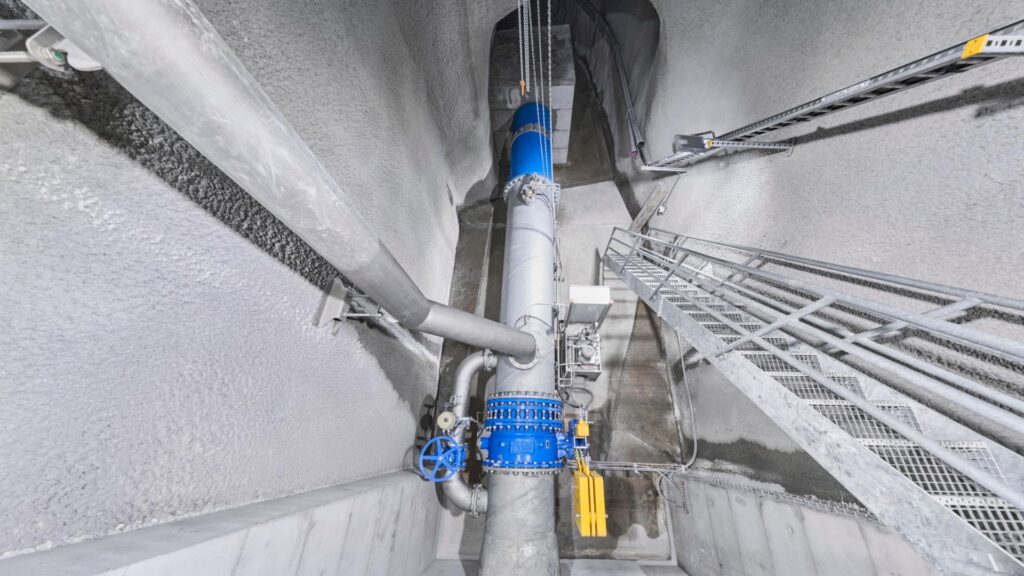Apparatekammer im WKW Sousbach mit blauen Armaturen, Rohrleitungen und Metalltreppe in einem Betontunnel.