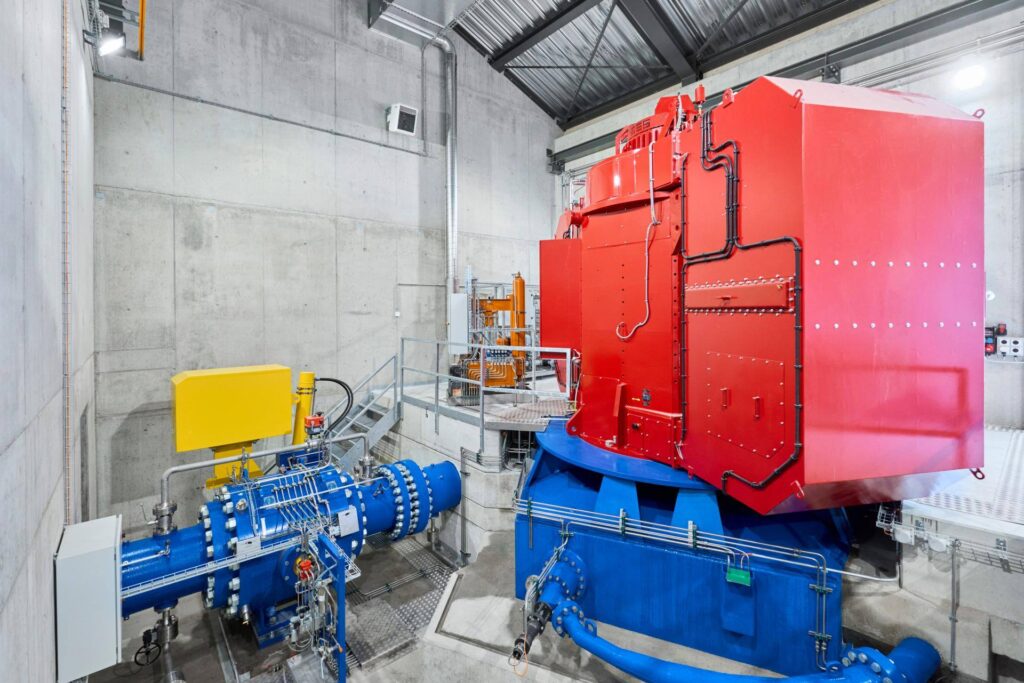 Roter Generator und blaue Maschinenausrüstung im Wasserkraftwerk Sousbach in einer modernen Industriehalle aus Sichtbeton.
