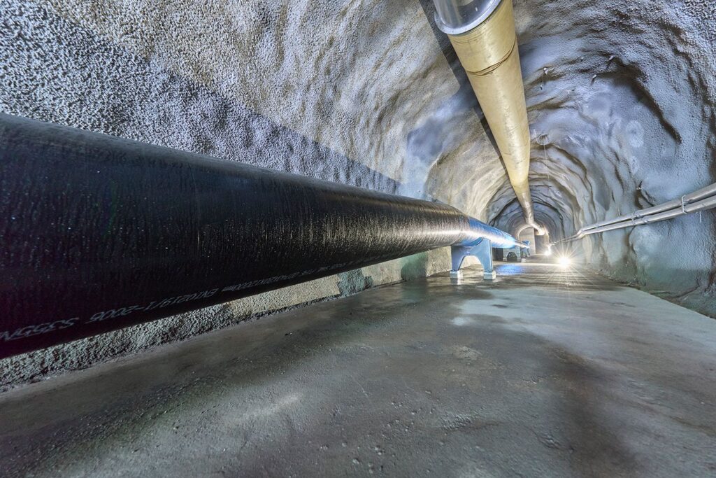 Schwarzes Stahlrohr im Sousbach Stollen des WKW, verlegt in einem betonierten Tunnel mit Belüftungssystem und Punktbeleuchtung.