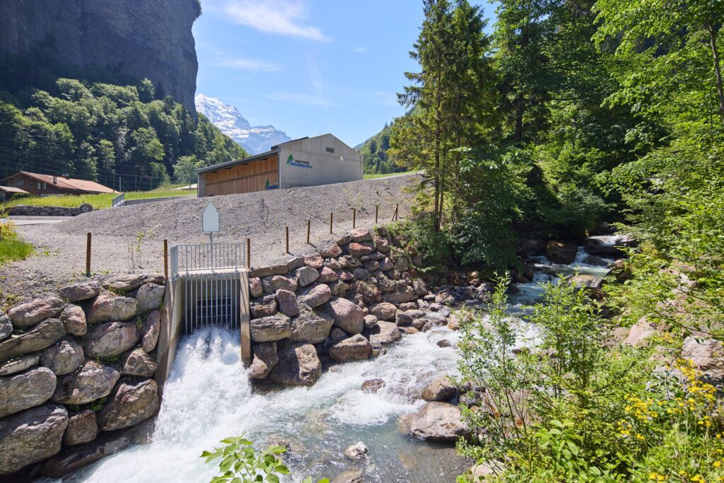 Wasserkraftwerk Sousbach: Unterwasserkanal mündet in einen Gebirgsbach vor bewaldeten Bergen und schneebedeckten Gipfeln.