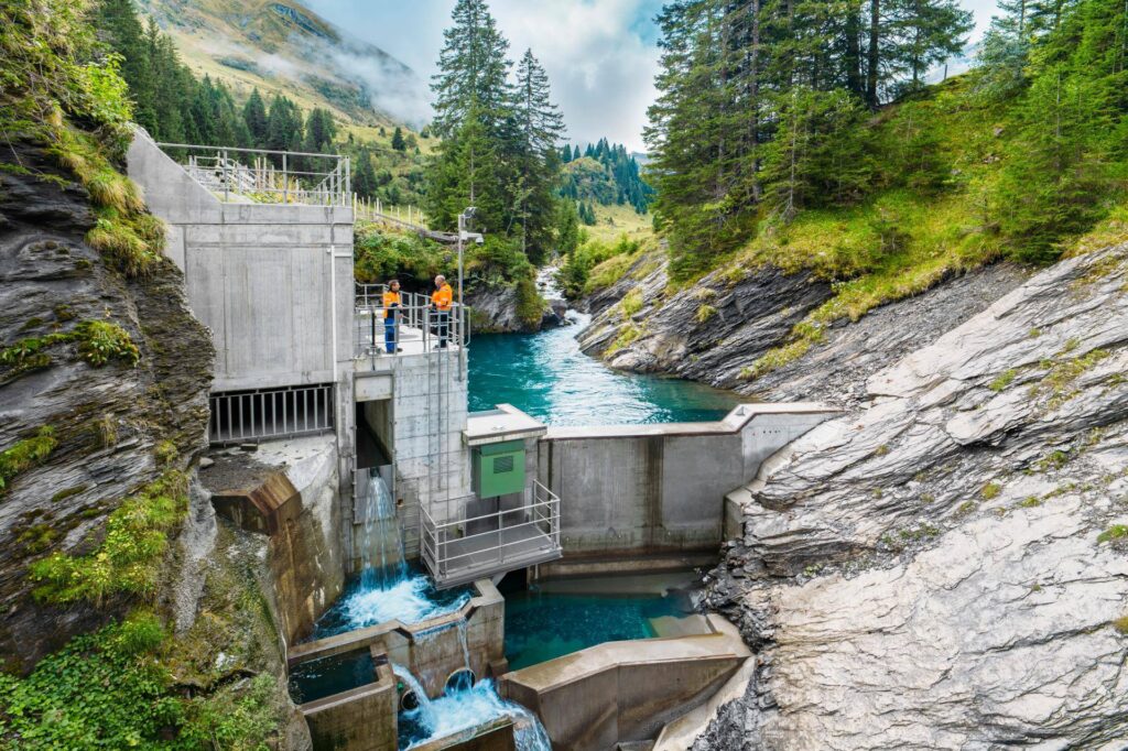 Wasserfassung am Sousbach: Mitarbeiter in Warnwesten auf der Betonstruktur des Wasserkraftwerks in alpiner Landschaft.