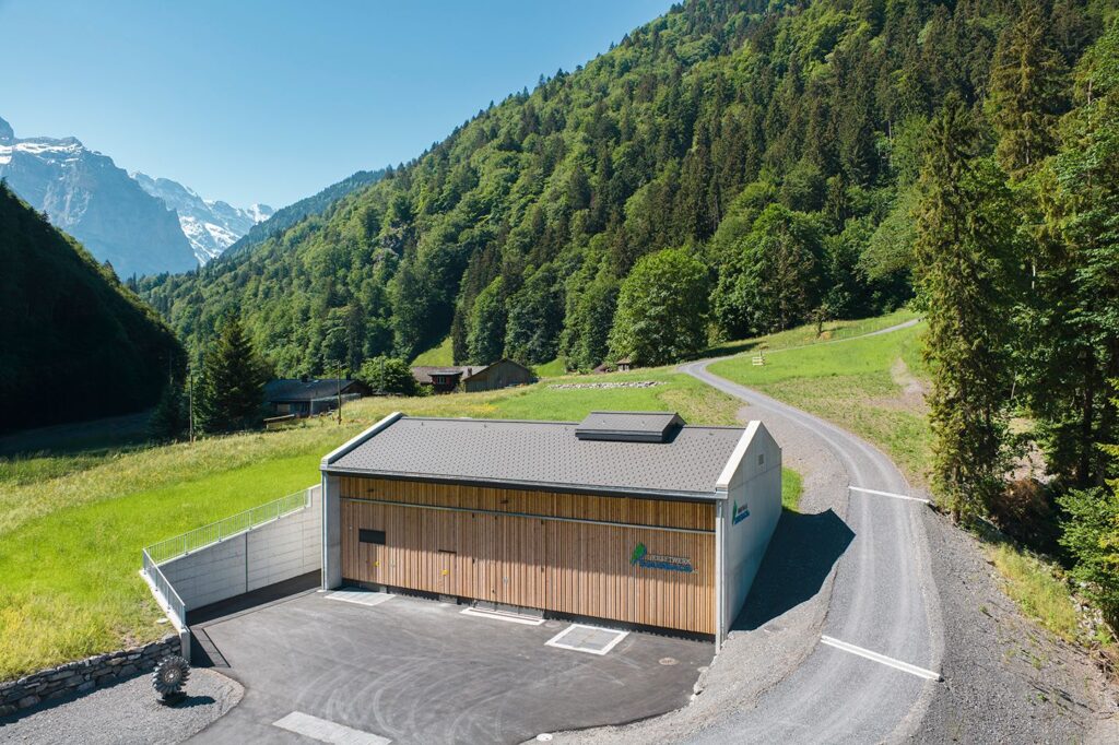Zentralengebäude des Wasserkraftwerks Sousbach mit Holzfassade vor bewaldetem Berghang und schneebedeckten Gipfeln.