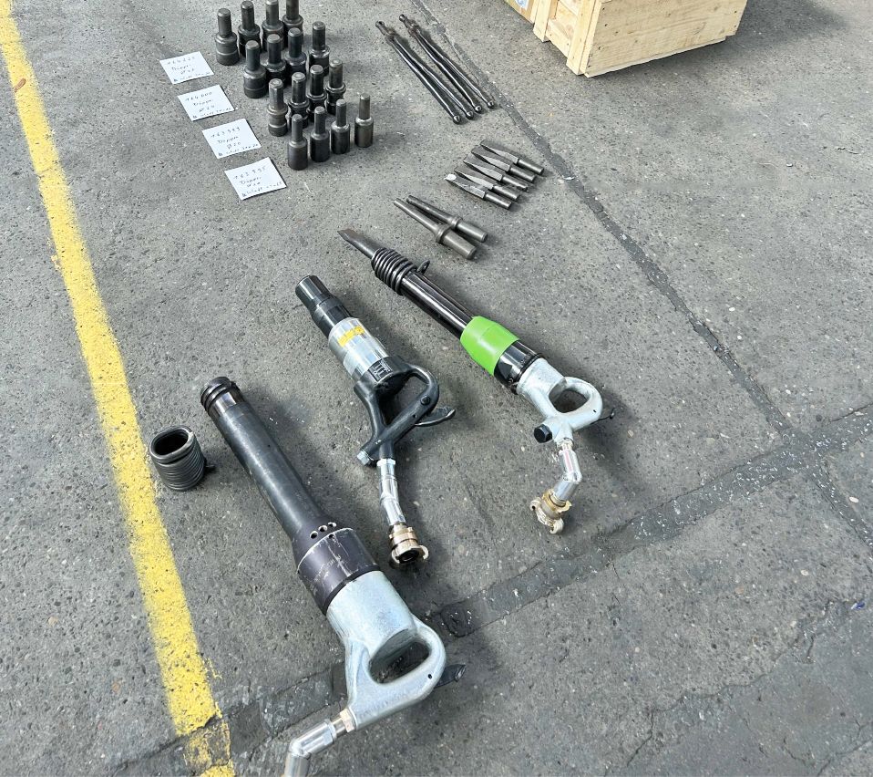 Druckluft-Werkzeuge wie Meißelhämmer, Bohrer und Zubehör geordnet auf Werkstattboden neben einer gelben Markierungslinie.