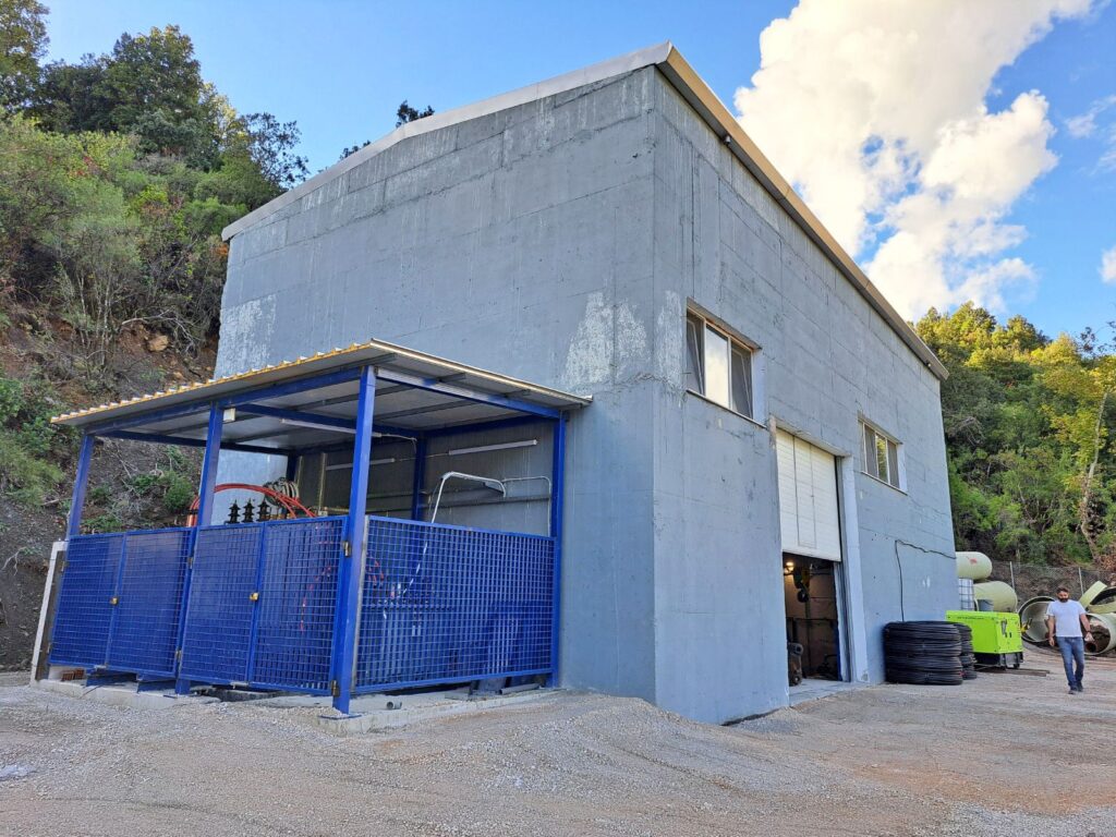 Industriegebäude aus grauem Beton mit blau eingezäunter Trafostation und offenem Rolltor vor bewaldetem Hang.