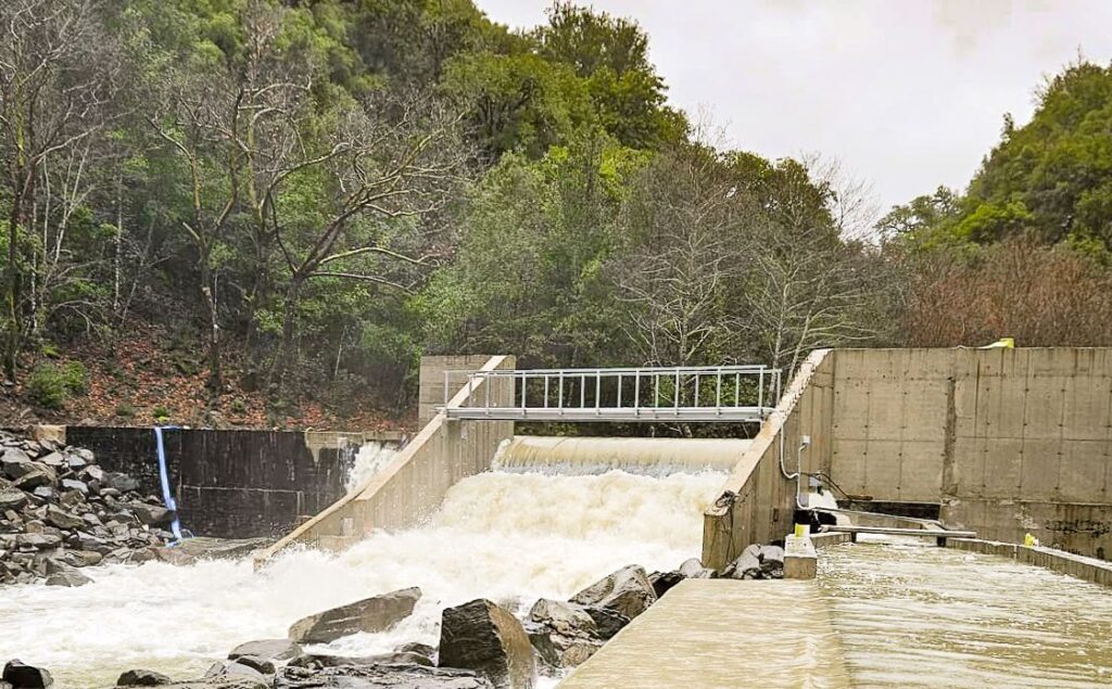 Staudamm aus Beton mit überlaufendem Wasserfall in einem Waldgebiet, Metallsteg über dem Wehr und felsiges Flussbett.