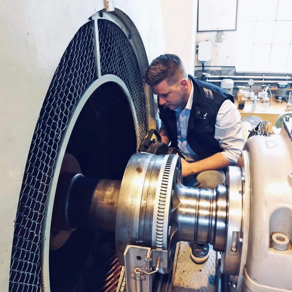 Techniker prüft mit einem Messgerät die Antriebswelle einer großen Industriemaschine in einer Werkstatt.