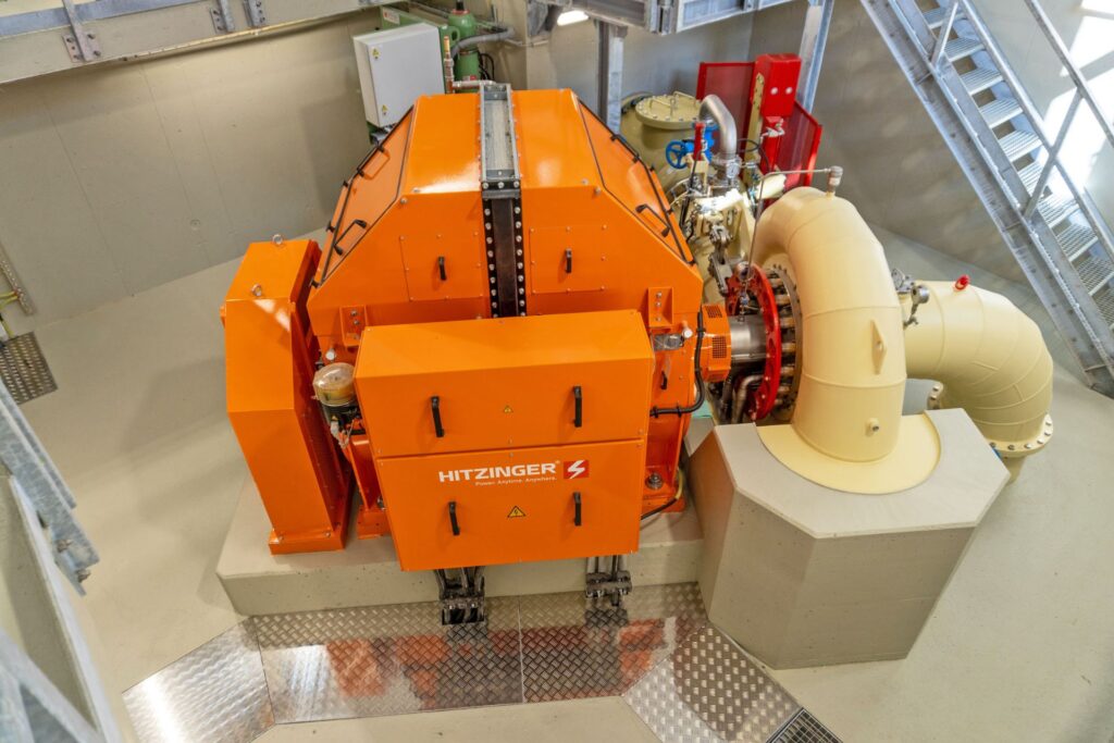 Draufsicht auf eine orangefarbene Hitzinger Maschine im Kraftwerk Hüttenberg mit großen Rohrleitungen und Treppenaufgang.